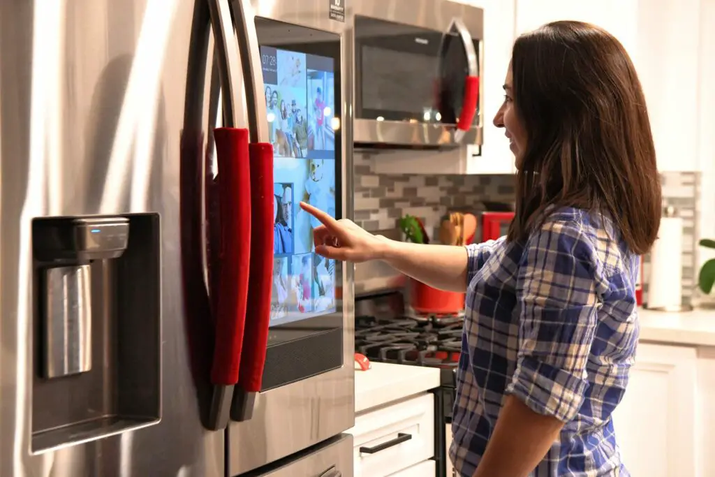 A Woman Demonstrates The Smart Capabilities Of Her Refrigerator By Pointing At A Screen.